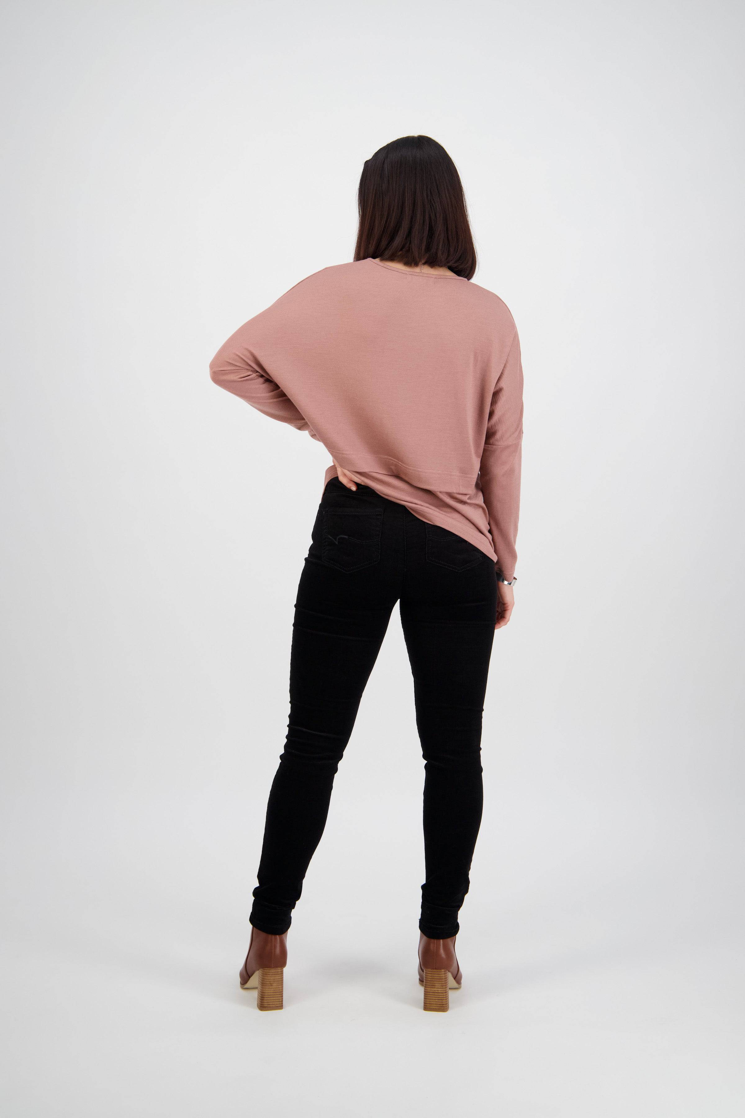 A woman with shoulder-length dark hair stands facing away, wearing a loose pink long-sleeve top, Vassalli Origin Cord Pull On pants in black, and tan heeled shoes against a plain white background.
