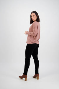 A woman with shoulder-length dark hair, in a loose mauve top and Vassalli Origin Cord Pull On pants in black, paired with brown heeled boots, stands in a white studio, turned slightly to the side and looking at the camera.