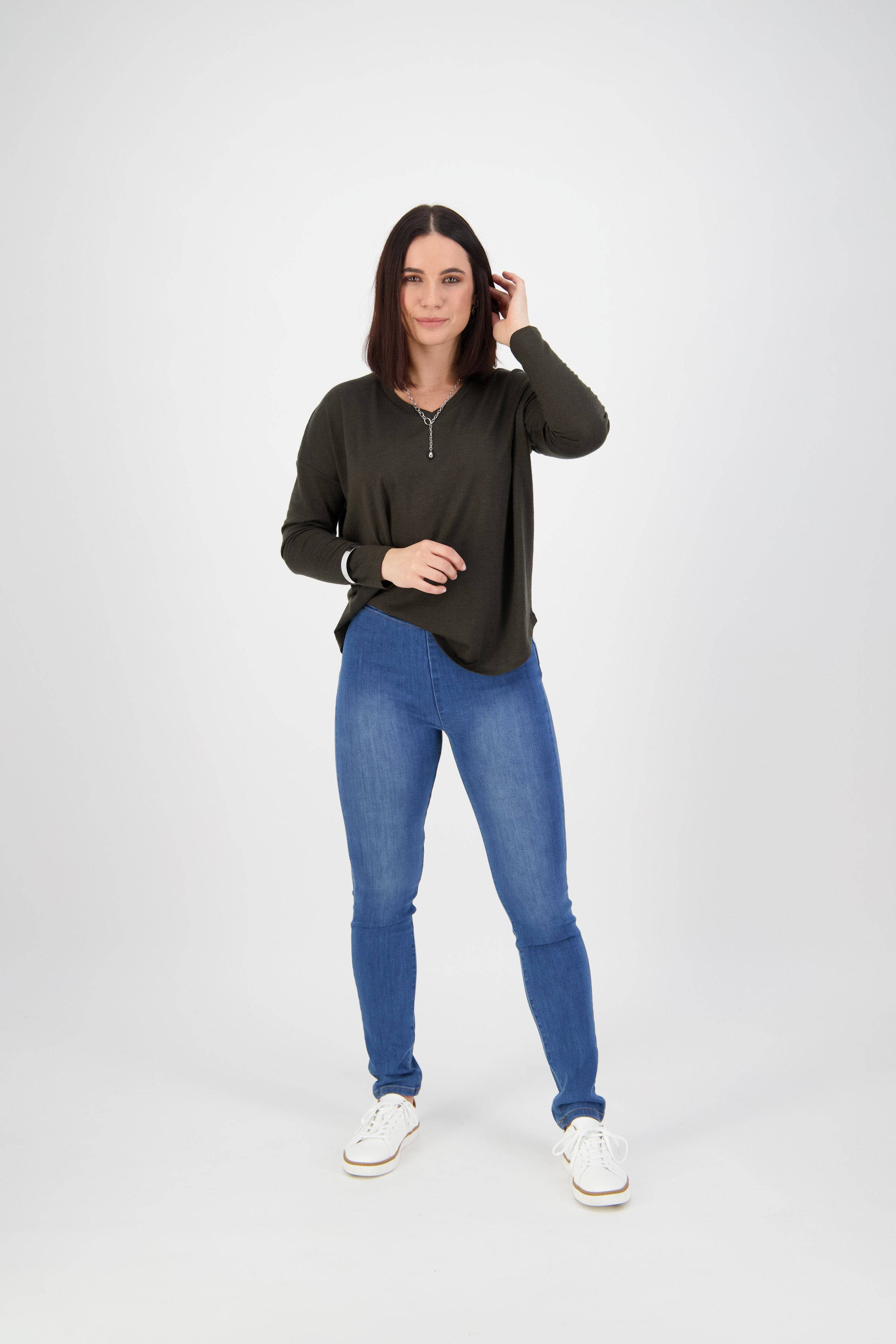 A woman with straight dark hair stands on a white background, wearing a dark green long-sleeve top, Vassalli Origin Pull On - New Blue jeans in stretch denim, and white sneakers. She looks at the camera, one hand touching her hair.
