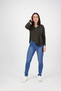 A woman with straight dark hair wears Vassalli's Origin Pull On - New Blue skinny leg jeans, a dark green long-sleeve top, and white sneakers. She stands against a plain white background, touching her hair with a neutral expression.