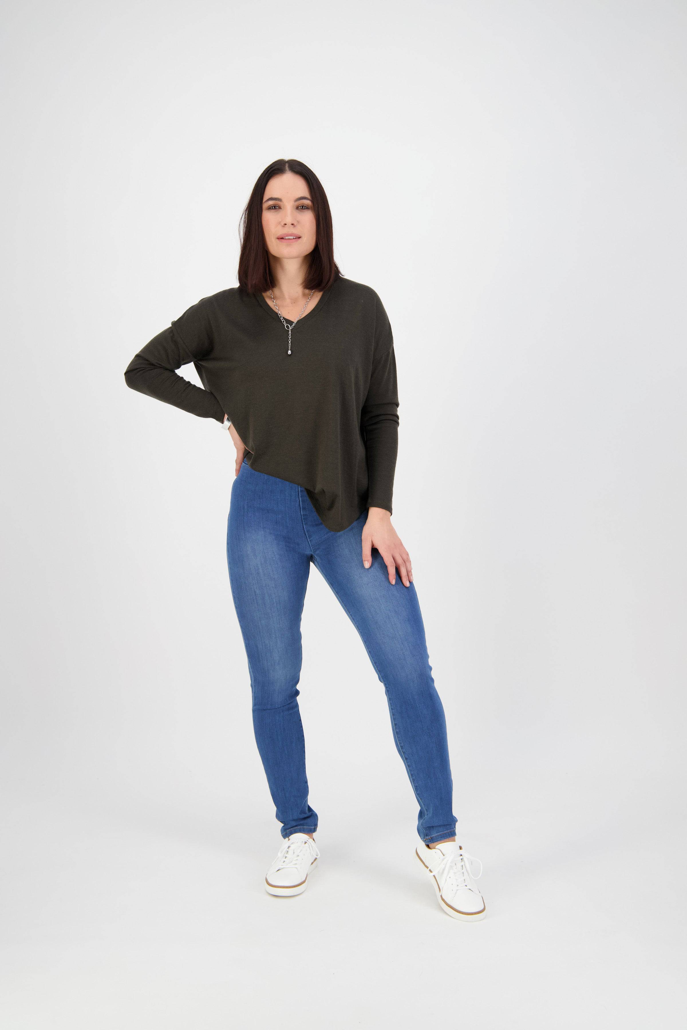 A woman poses against a plain white background, wearing a dark long-sleeve top, Vassalli Origin Pull On jeans in New Blue stretch denim, and white sneakers. Her straight shoulder-length hair frames her face as she rests one hand on her hip.