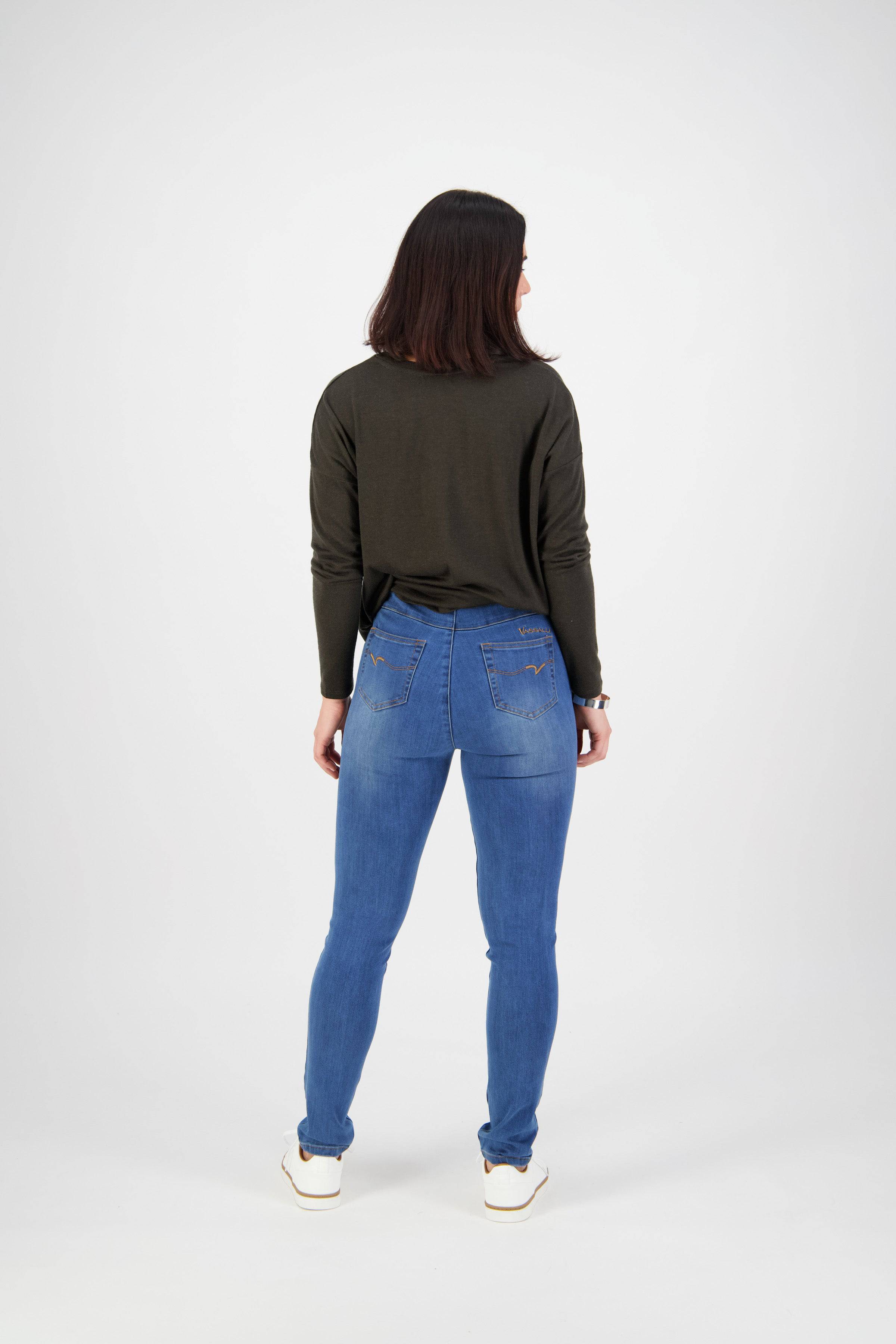 A person with shoulder-length dark hair, wearing a long-sleeve olive green top, Vassalli Origin Pull On - New Blue jeans, and white shoes stands facing away from the camera against a plain white background.