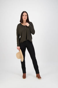 A woman with straight brown hair poses against a white background, wearing a dark green long-sleeve top, Vassalli Bay Ponti Pull On pants in black, brown ankle boots, and holding a beige hat while looking confidently at the camera.