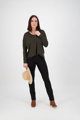 A woman with straight brown hair poses against a white background, wearing a dark green long-sleeve top, Vassalli Bay Ponti Pull On pants in black, brown ankle boots, and holding a beige hat while looking confidently at the camera.