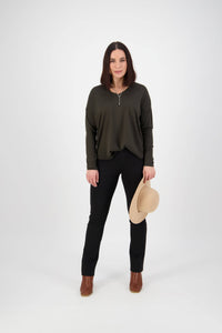 A woman faces forward against a white background, wearing a dark green long-sleeve top and Vassalli Bay Ponti Pull On black slim leg pants made from stretch fabric, brown boots, and holding a beige hat in her left hand.