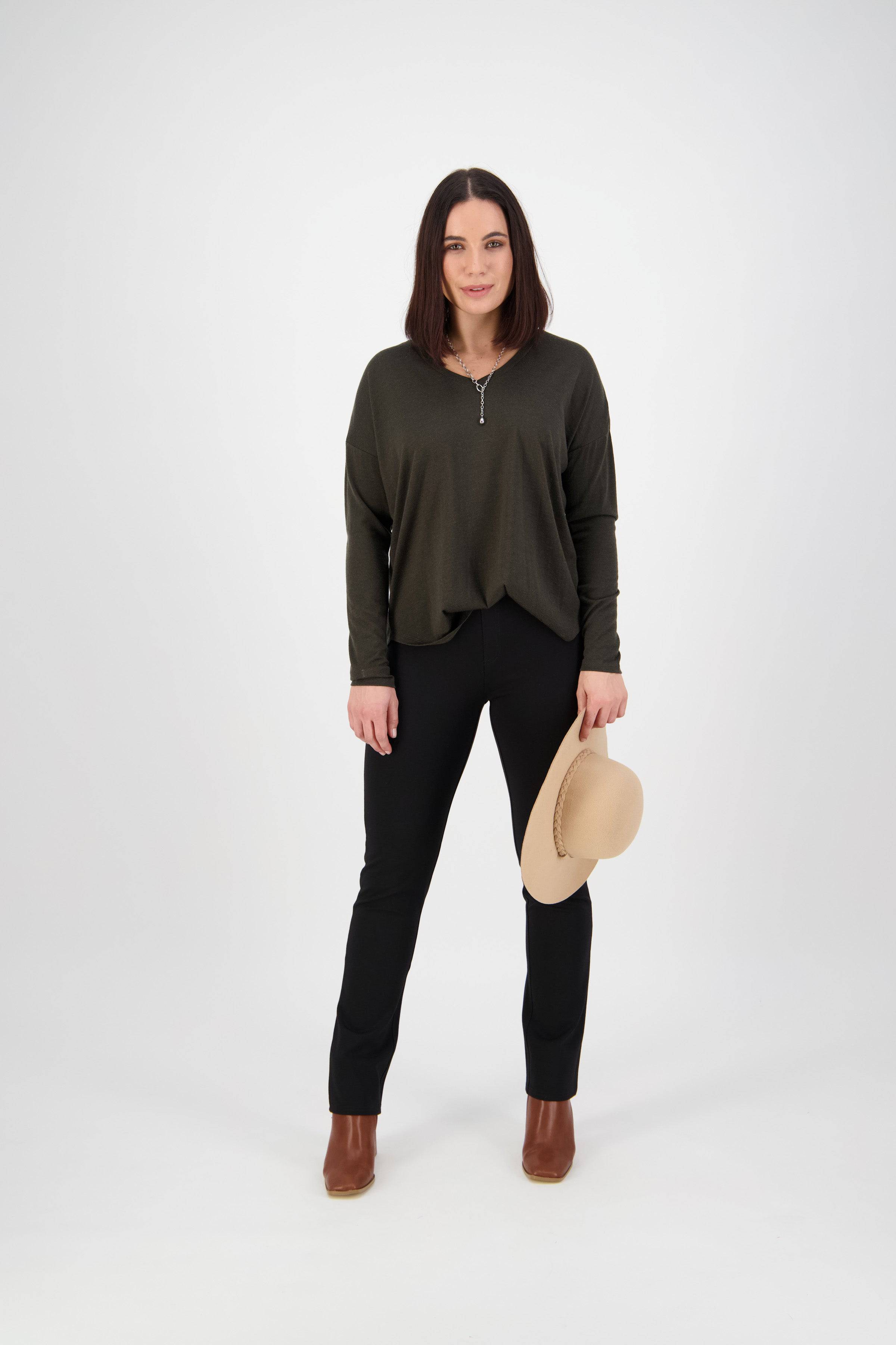 A woman faces forward against a white background, wearing a dark green long-sleeve top and Vassalli Bay Ponti Pull On black slim leg pants made from stretch fabric, brown boots, and holding a beige hat in her left hand.
