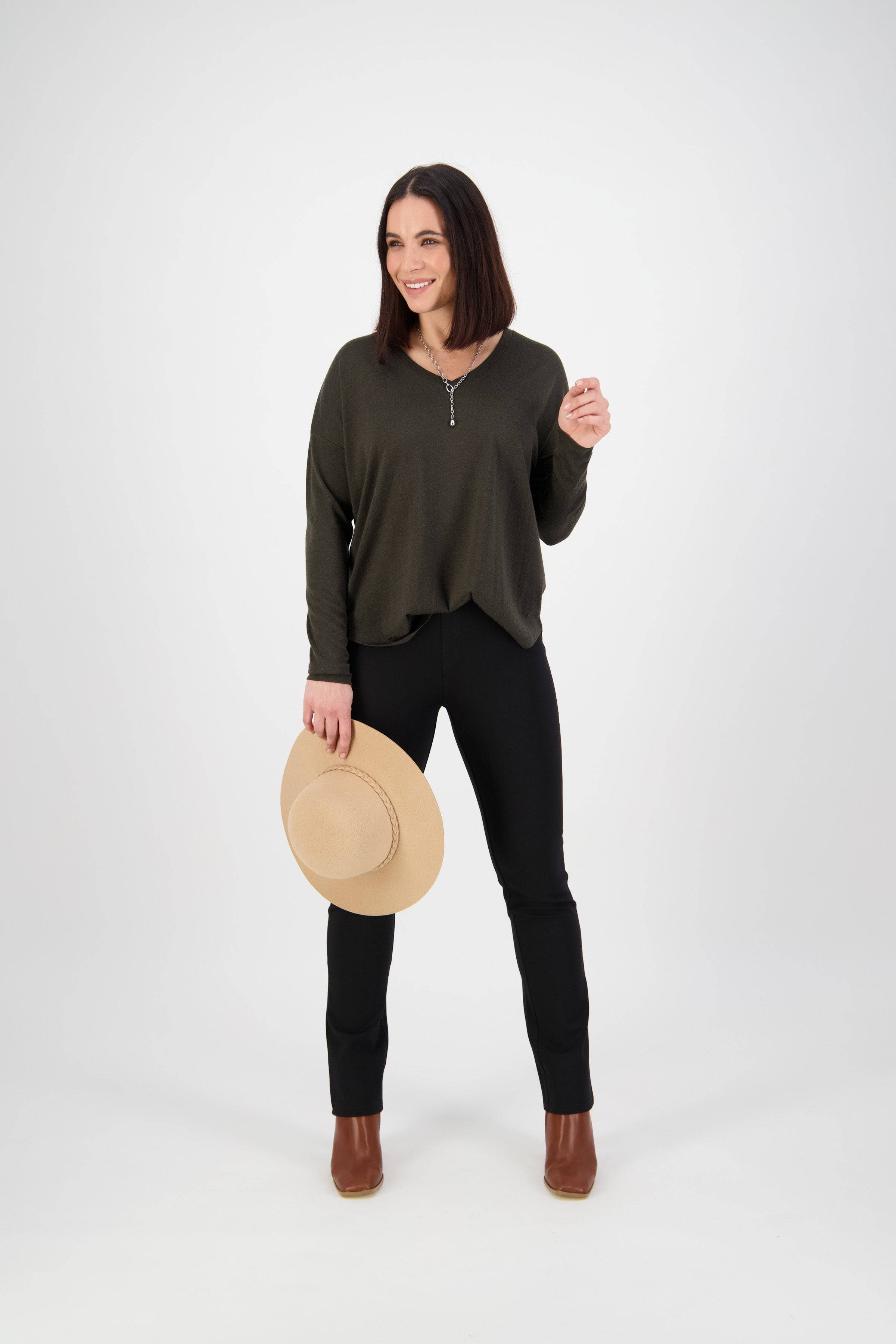 A woman with shoulder-length dark hair wears Vassalli’s Bay Ponti Pull On - Black pants, a dark green long-sleeve top, and brown ankle boots. She smiles while holding a beige hat in her left hand against a white background.