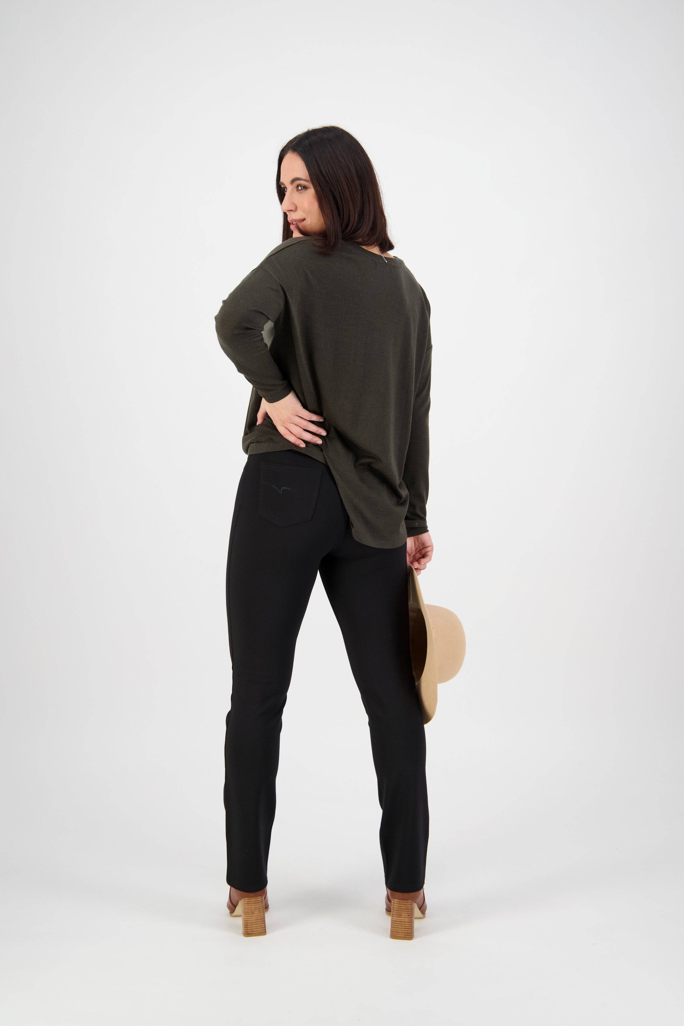 A woman faces away, looking over her shoulder, wearing Vassalli's Bay Ponti Pull On - Black slim leg pants with a dark green long-sleeve top and heels, holding a beige hat in her left hand against a white background.