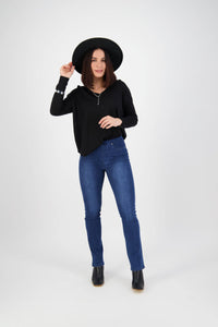 A woman smiles against a white background, touching her black hat while wearing a black long-sleeve top, black boots, and Vassalli Bay Pull On - Blue Denim (With Front Fade) slim leg jeans.
