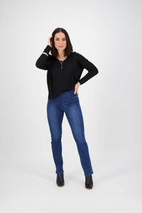 A woman stands in a studio with a white background, wearing Vassalli Bay Pull On - Blue Denim (With Front Fade) jeans, a black long-sleeve top, and black ankle boots. She has shoulder-length dark hair and touches her head while smiling slightly.