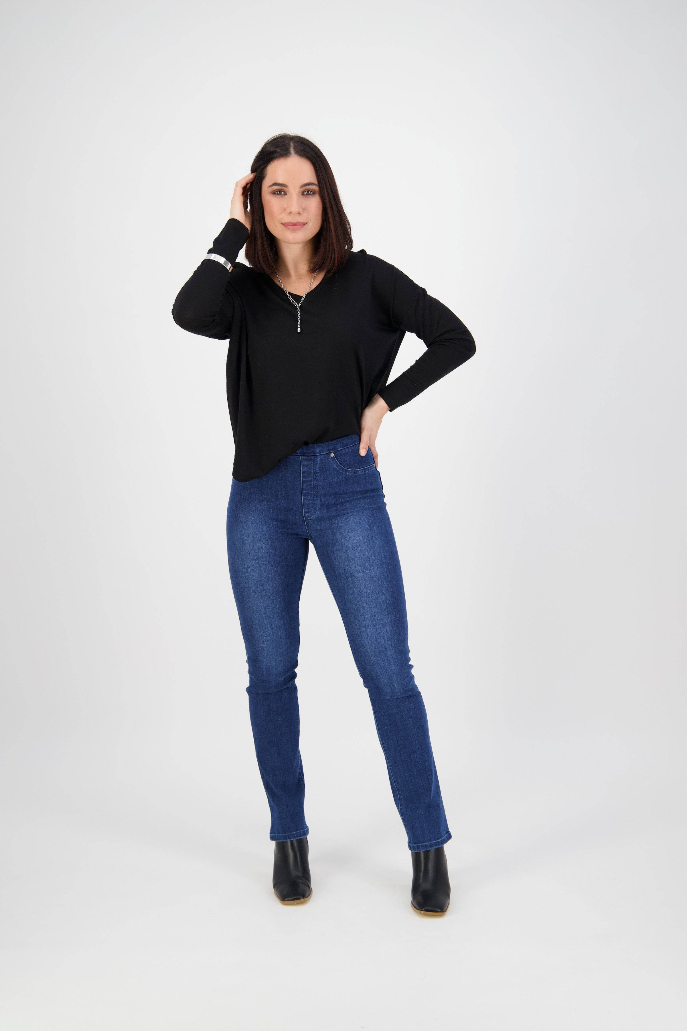 A woman stands in a studio with a white background, wearing Vassalli Bay Pull On - Blue Denim (With Front Fade) jeans, a black long-sleeve top, and black ankle boots. She has shoulder-length dark hair and touches her head while smiling slightly.