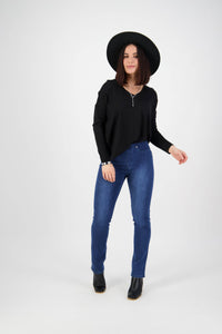 A woman wearing Vassalli's Bay Pull On - Blue Denim (With Front Fade) jeans stands against a white background, paired with a black wide-brim hat, black long-sleeve top, and black ankle boots, posing relaxedly with a slight smile.