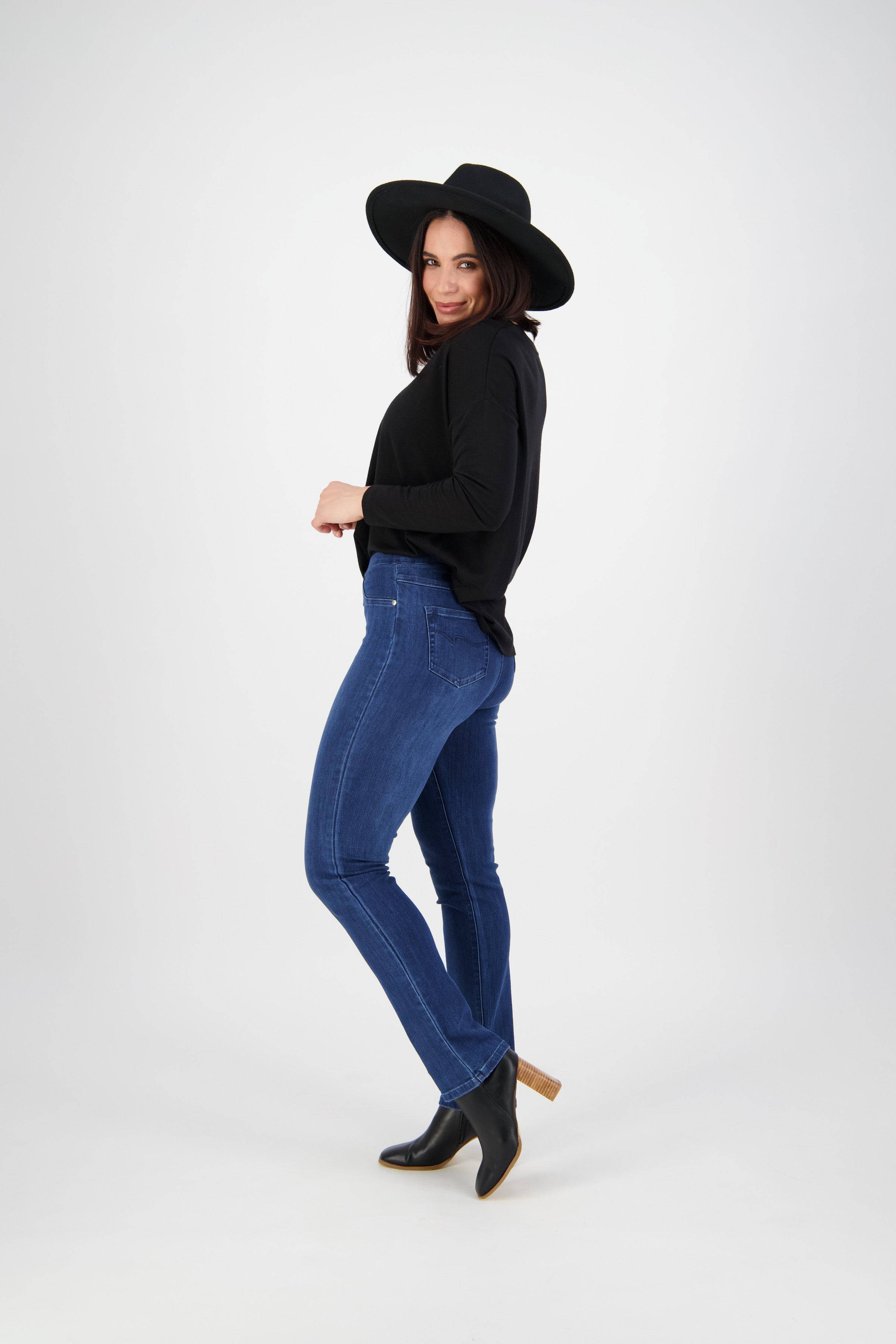 A woman poses sideways against a white background, smiling slightly with one foot forward, wearing Vassalli's Bay Pull On - Blue Denim (With Front Fade) jeans paired with a black wide-brimmed hat, long-sleeve top, and ankle boots.