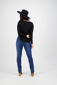 A person in Vassalli Bay Pull On - Blue Denim (With Front Fade) jeans, a black long-sleeve top, black hat, and heeled boots stands facing away with one arm behind their back against a plain white background.