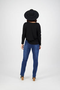 A person stands facing away from the camera against a plain white background, wearing Vassalli Bay Pull On - Blue Denim (With Front Fade) jeans, a black wide-brimmed hat, a black long-sleeve top, and heeled boots.