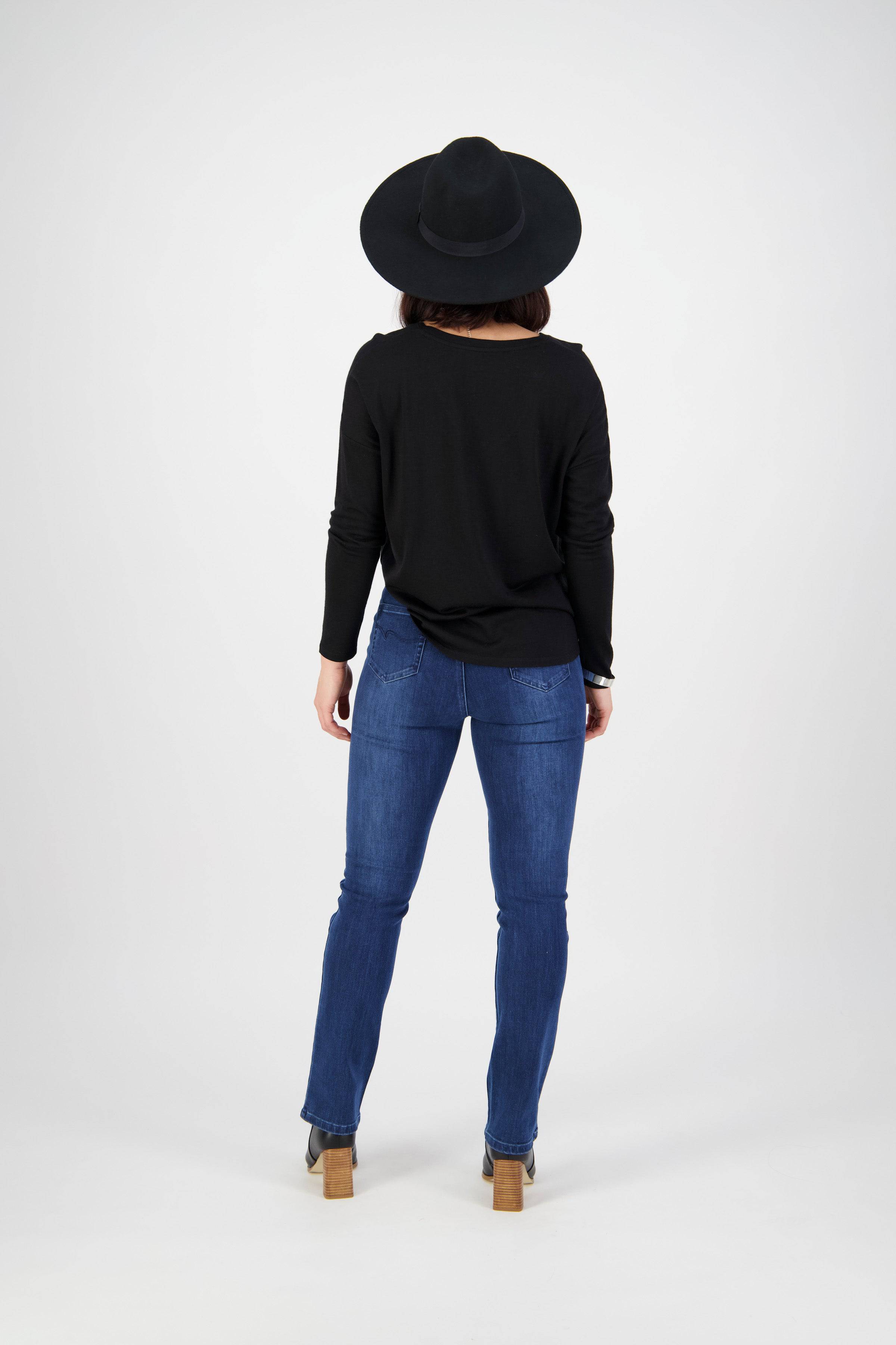 A person stands facing away from the camera against a plain white background, wearing Vassalli Bay Pull On - Blue Denim (With Front Fade) jeans, a black wide-brimmed hat, a black long-sleeve top, and heeled boots.