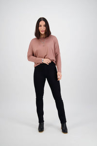 A woman with straight, shoulder-length brown hair faces forward, wearing a mauve long-sleeve top, Vassalli's Bay Pull On - Black Denim pants, and black ankle boots against a plain light background.