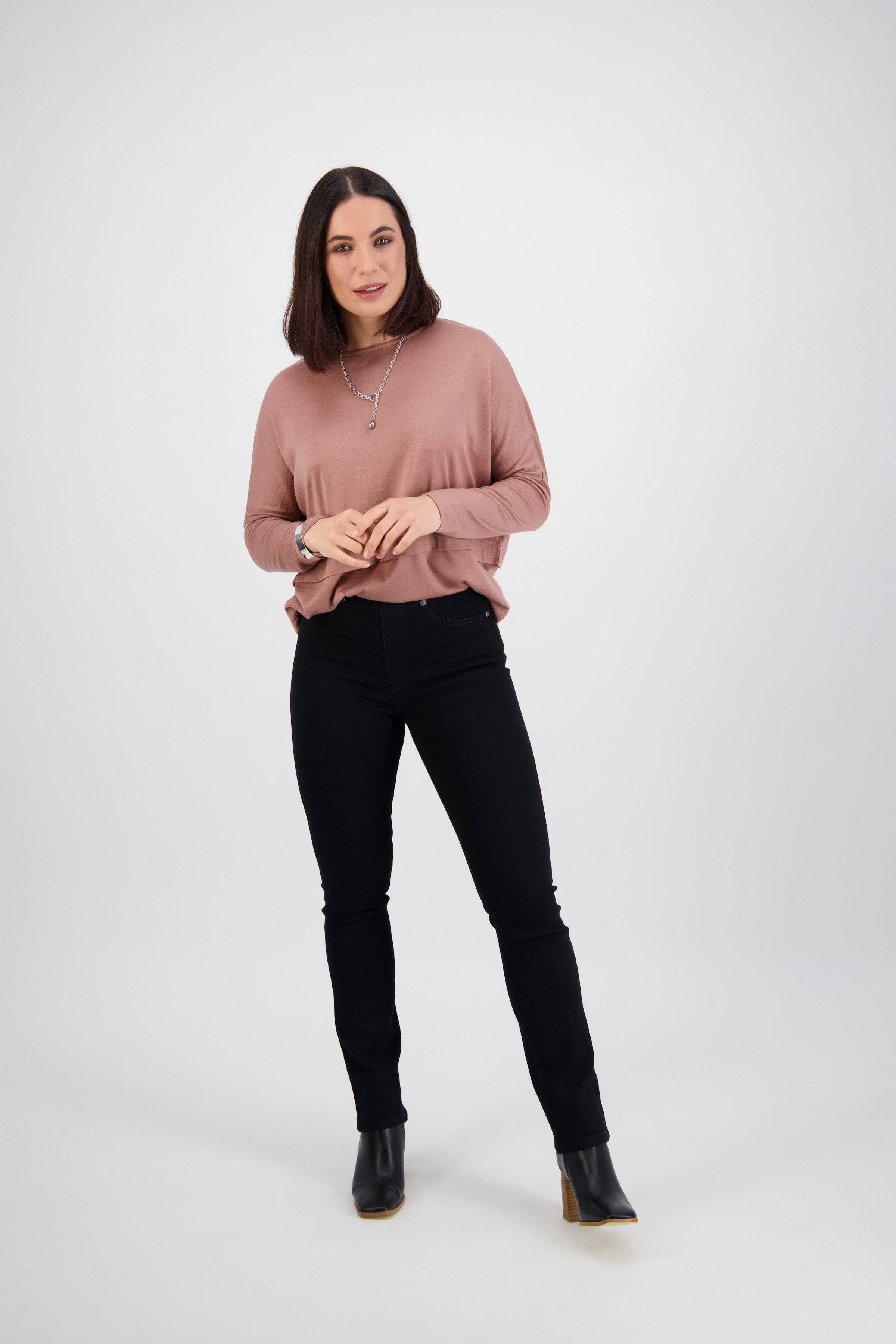 A woman with shoulder-length dark hair, in a pink long-sleeve top and Vassalli Bay Pull On - Black Denim pants, stands against a white background wearing black ankle boots, looking forward with a relaxed posture and slight smile.