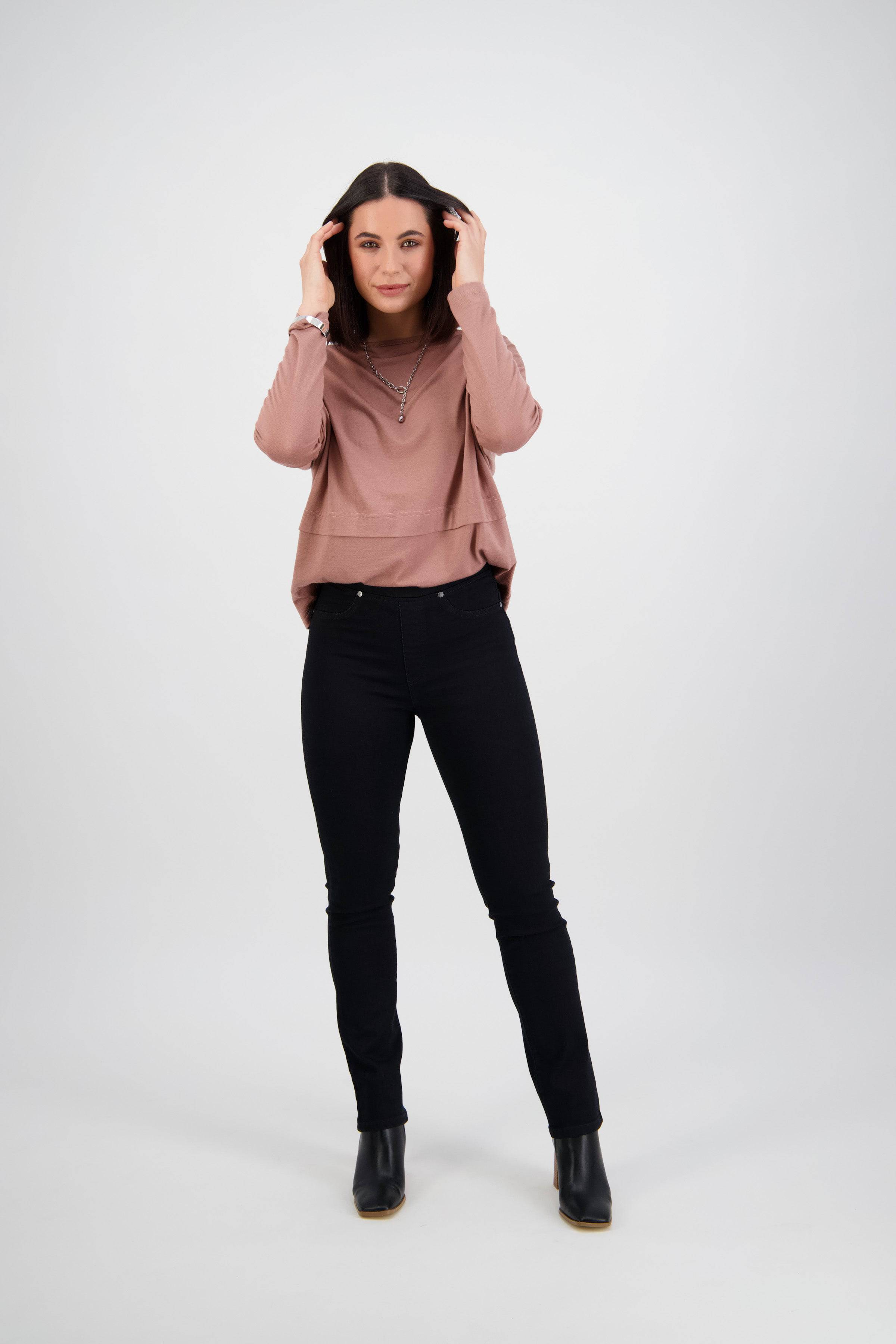 A woman with long dark hair smiles confidently as she holds her hair, wearing a mauve long-sleeve top and Vassalli Bay Pull On - Black Denim jeans, paired with black ankle boots against a plain light background.
