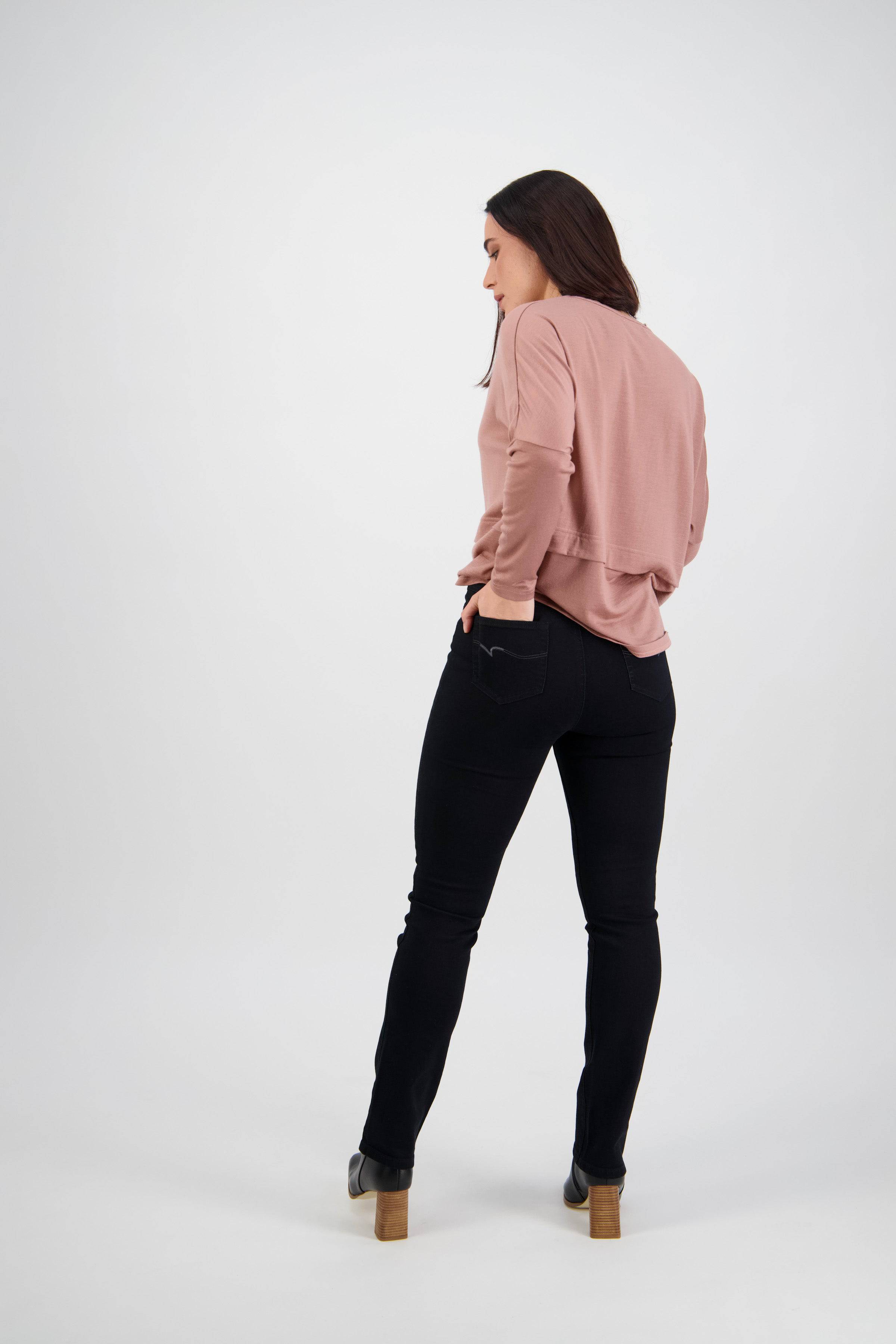 A woman faces away with her head turned, wearing Vassalli's Bay Pull On - Black Denim pants, a pink long-sleeve top, and heeled ankle boots. She stands with her hands in her back pockets against a plain white background.