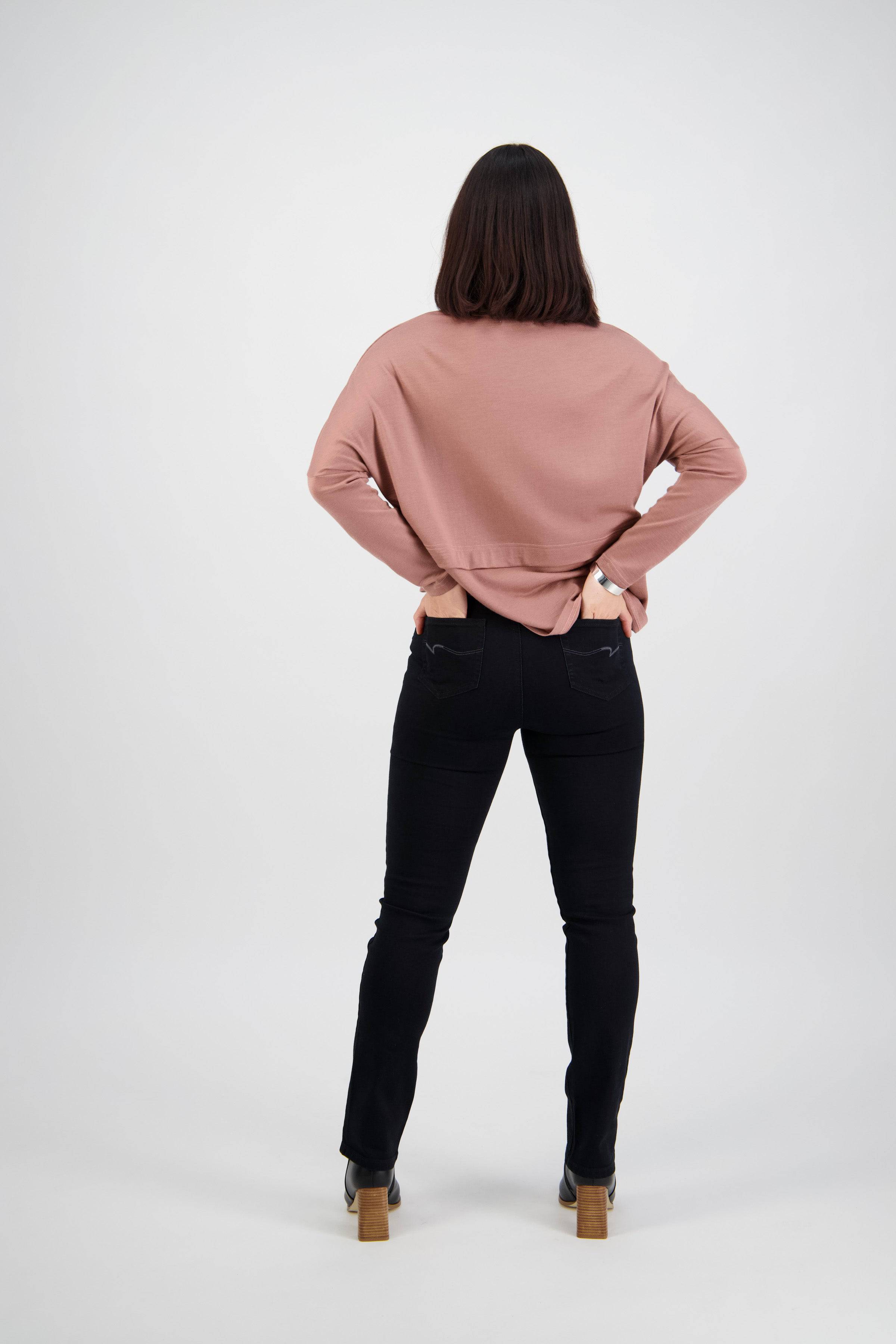A person with shoulder-length dark hair stands facing away, hands on hips, wearing a pink long-sleeve top, Vassalli Bay Pull On - Black Denim jeans, and heeled boots against a plain white background.