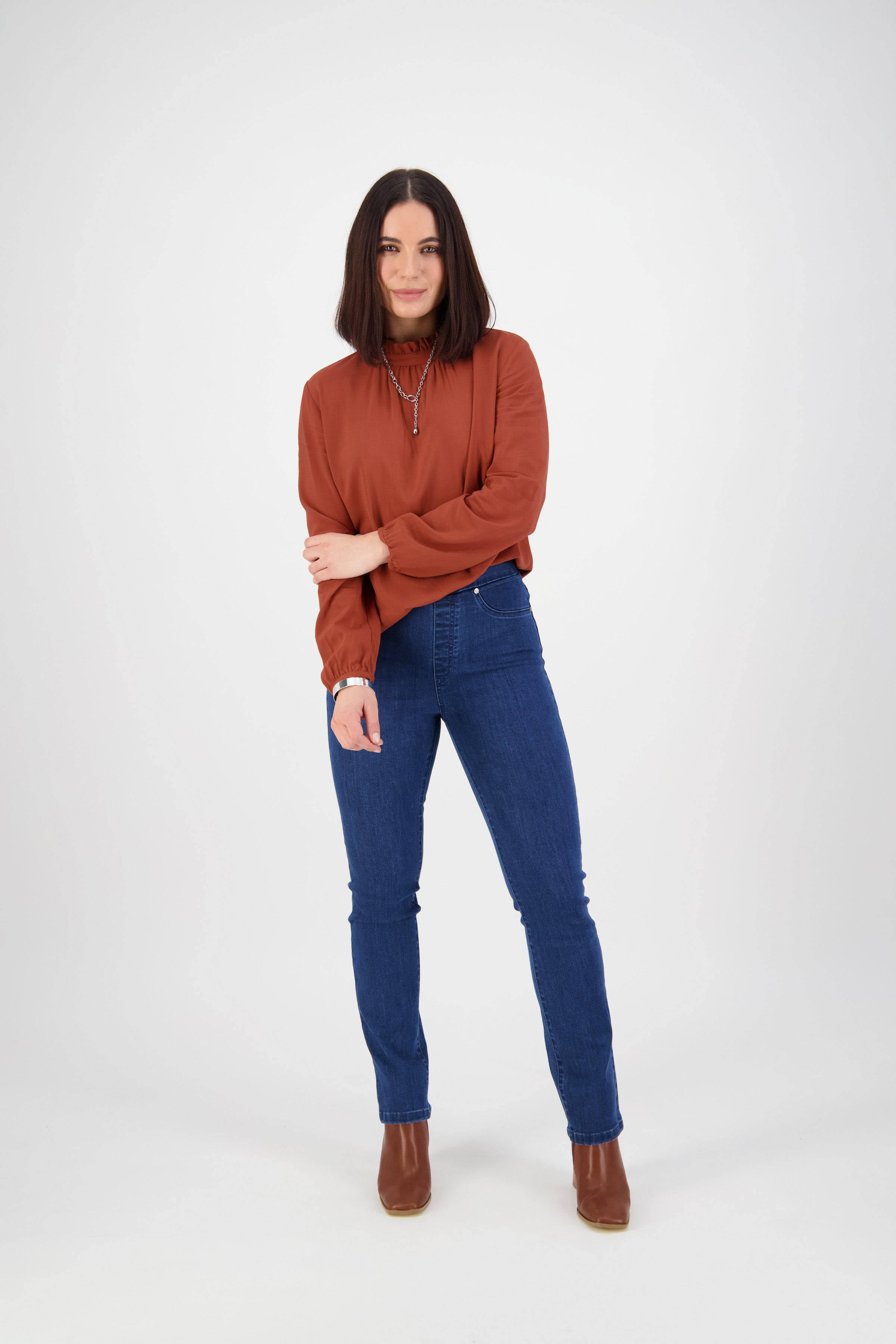 A woman with straight dark hair stands before a white background, wearing a rust blouse, brown ankle boots, and Vassalli's Bay Pull On - Blue Denim (Without Front Fade) jeans with a slim leg fit. She looks at the camera and smiles slightly.