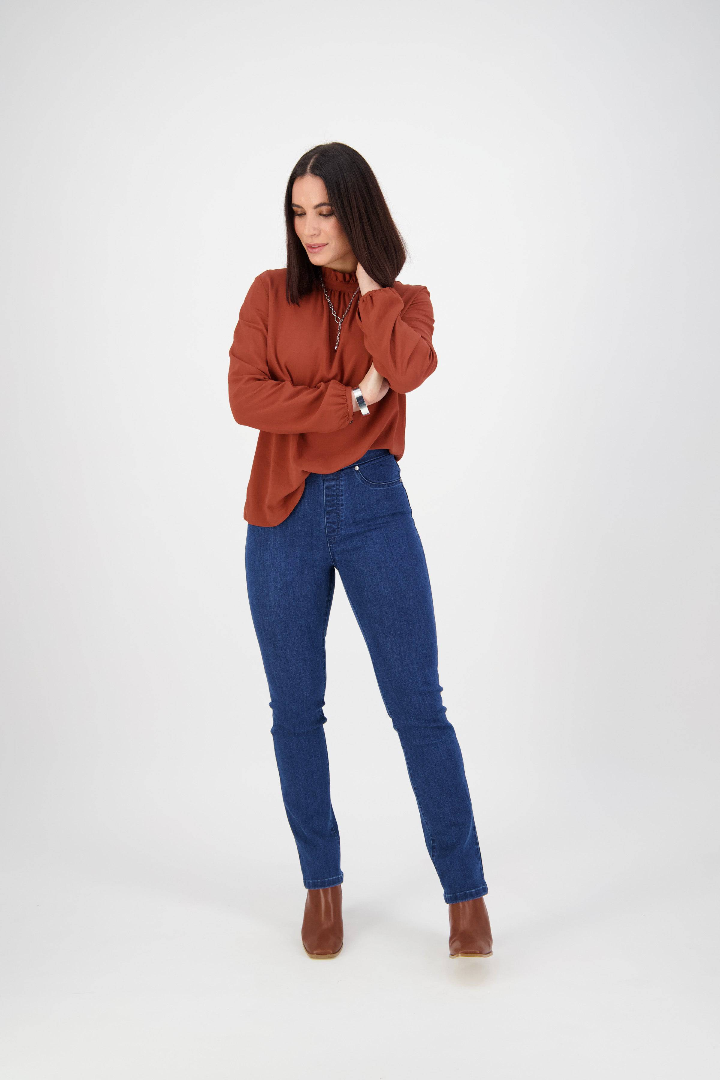 A woman with straight dark hair stands against a white background, arms crossed and looking down, wearing Vassalli's Bay Pull On - Blue Denim (Without Front Fade) jeans, a rust long-sleeve blouse, and brown ankle boots.