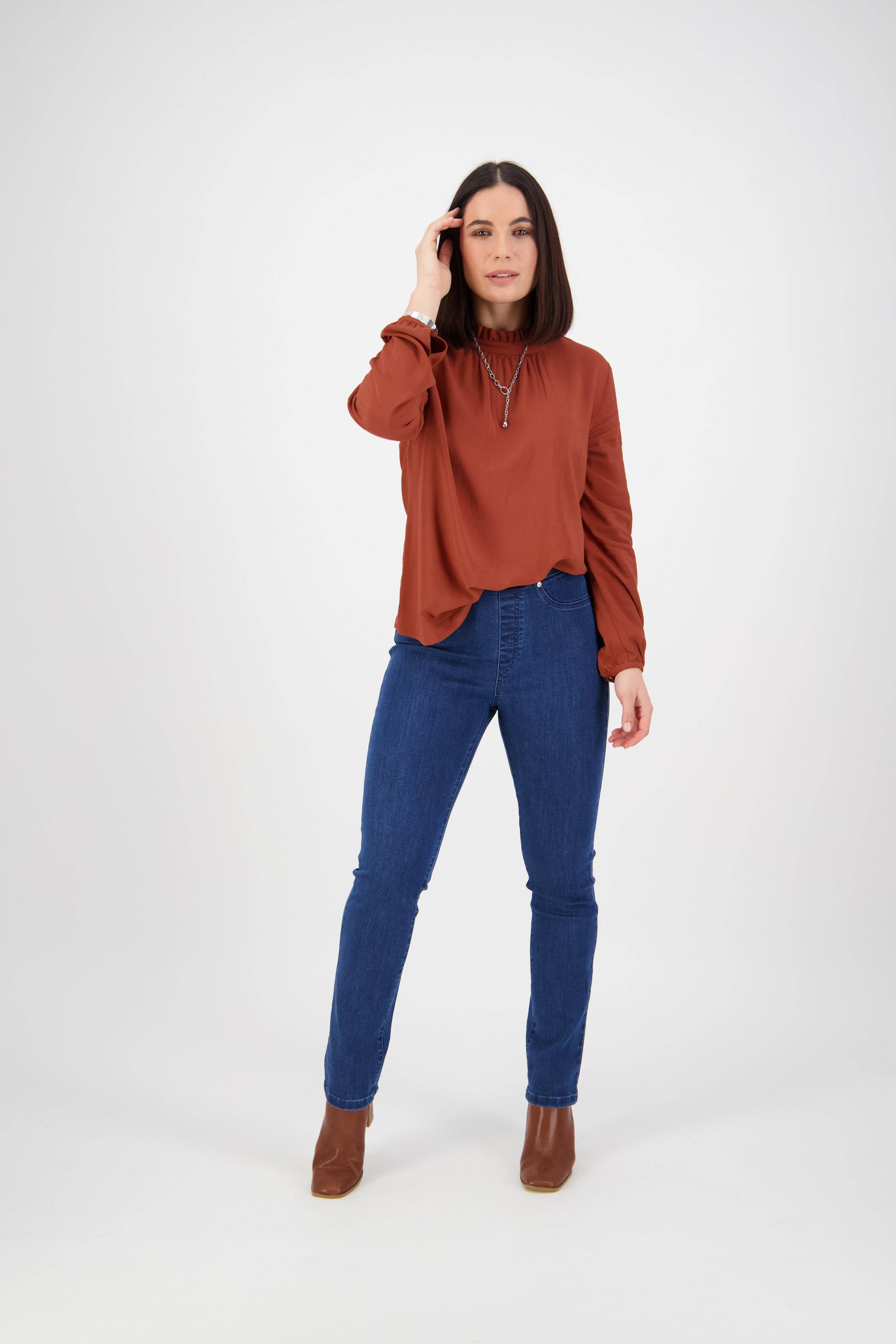 A woman stands facing forward against a plain white background, wearing Vassalli's Bay Pull On - Blue Denim (Without Front Fade) jeans, a rust long-sleeve blouse, and brown ankle boots, with her left hand touching her hair.