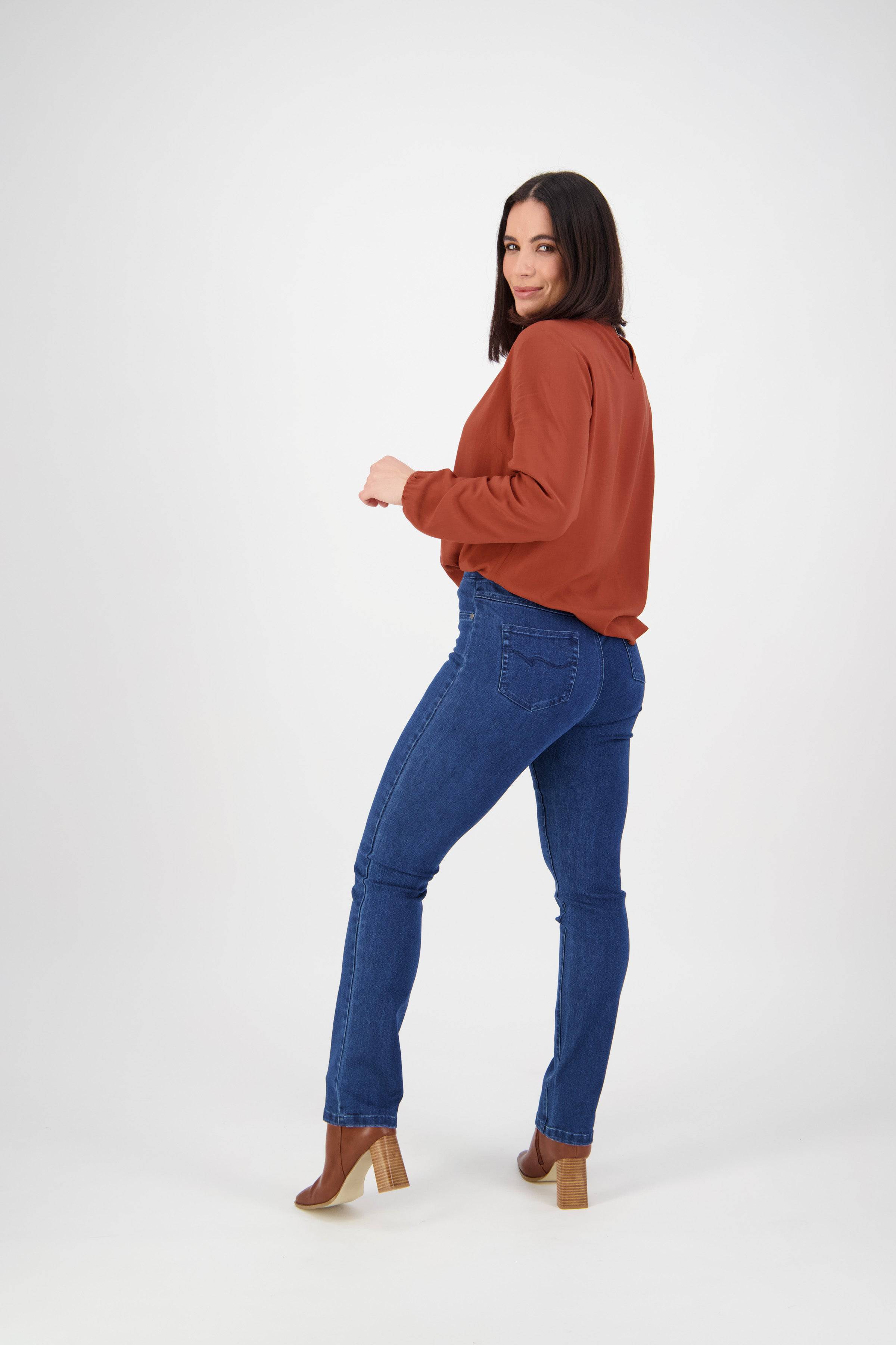 A woman with straight dark hair, in a rust long-sleeve top, Vassalli Bay Pull On blue denim jeans (no front fade), and heeled brown boots, stands in a white studio, looking over her shoulder and smiling slightly.