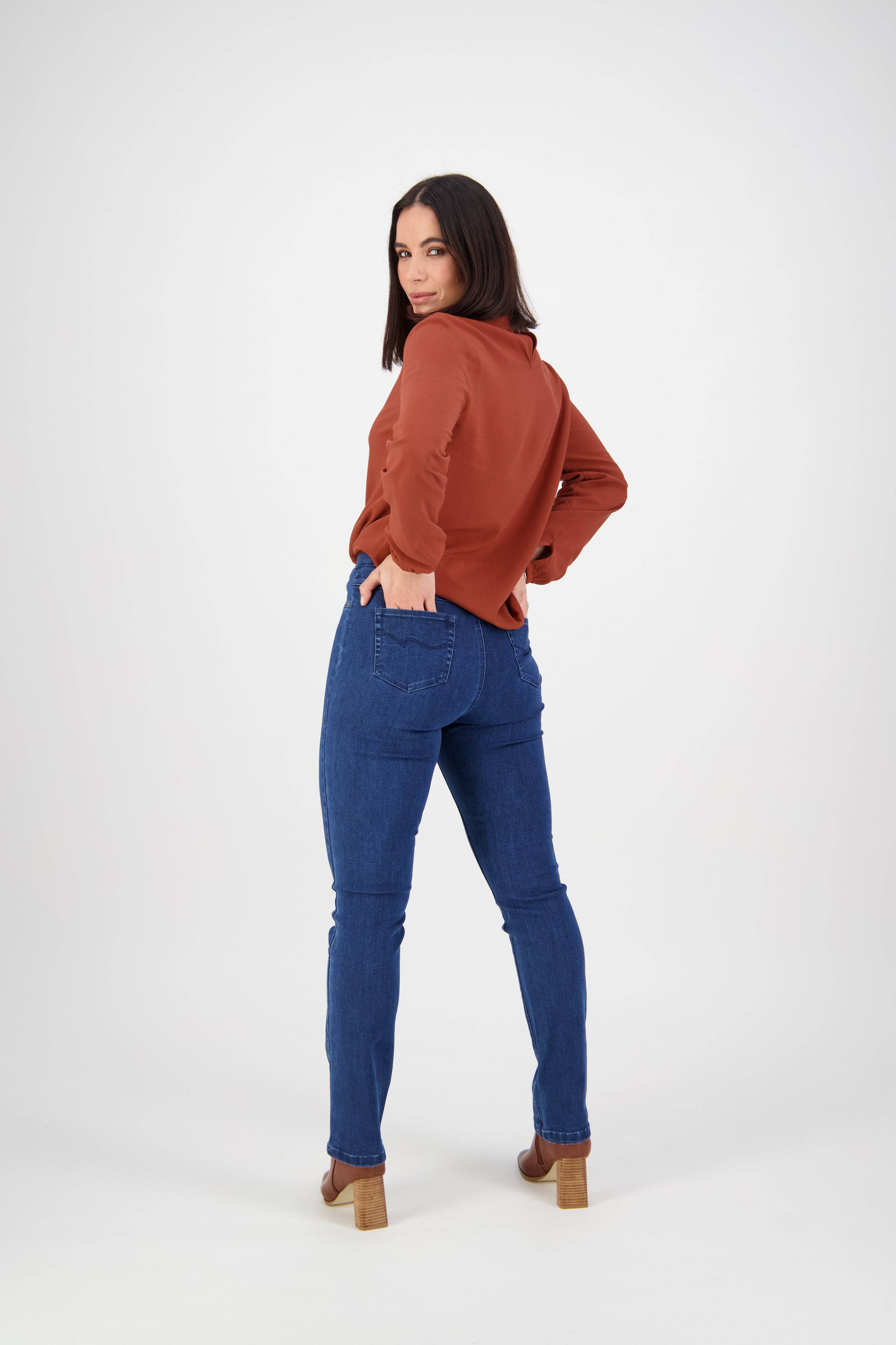 A woman poses with one hand on her hip, wearing a rust long-sleeve top, brown heeled shoes, and Vassalli Bay Pull On - Blue Denim (Without Front Fade) jeans, facing away but looking over her shoulder against a white background.
