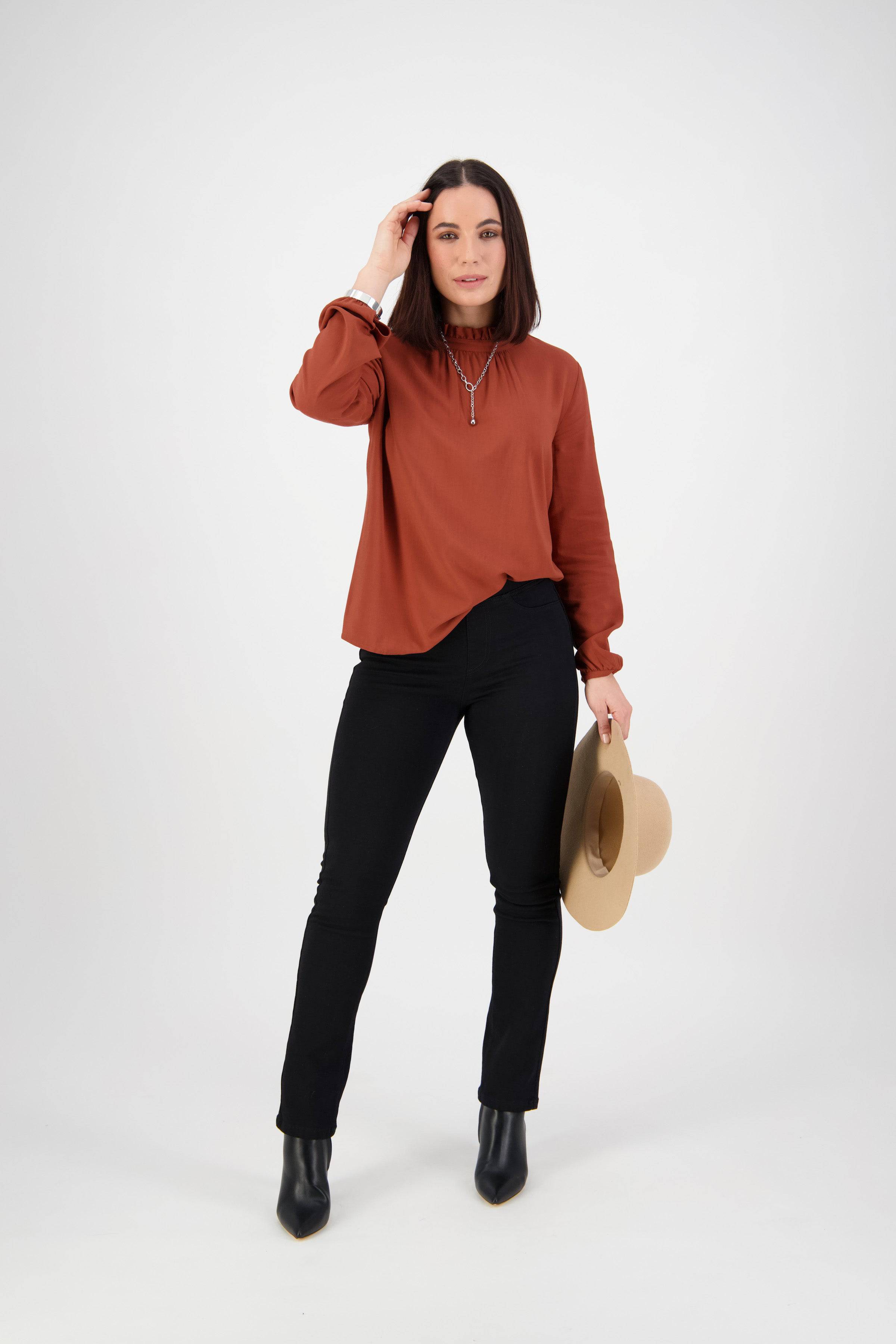 A woman stands against a white background, wearing a rust long-sleeve blouse and Vassalli Bay Knit Pull On pants in black with black boots. She holds a beige hat in one hand and touches her hair with the other.