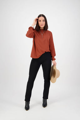 A woman stands against a white background, wearing a rust long-sleeve blouse and Vassalli Bay Knit Pull On pants in black with black boots. She holds a beige hat in one hand and touches her hair with the other.