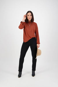 A woman stands against a plain white background, wearing a rust blouse, Vassalli Bay Knit Pull On - Black pants, black ankle boots, and holding a beige hat in her left hand.