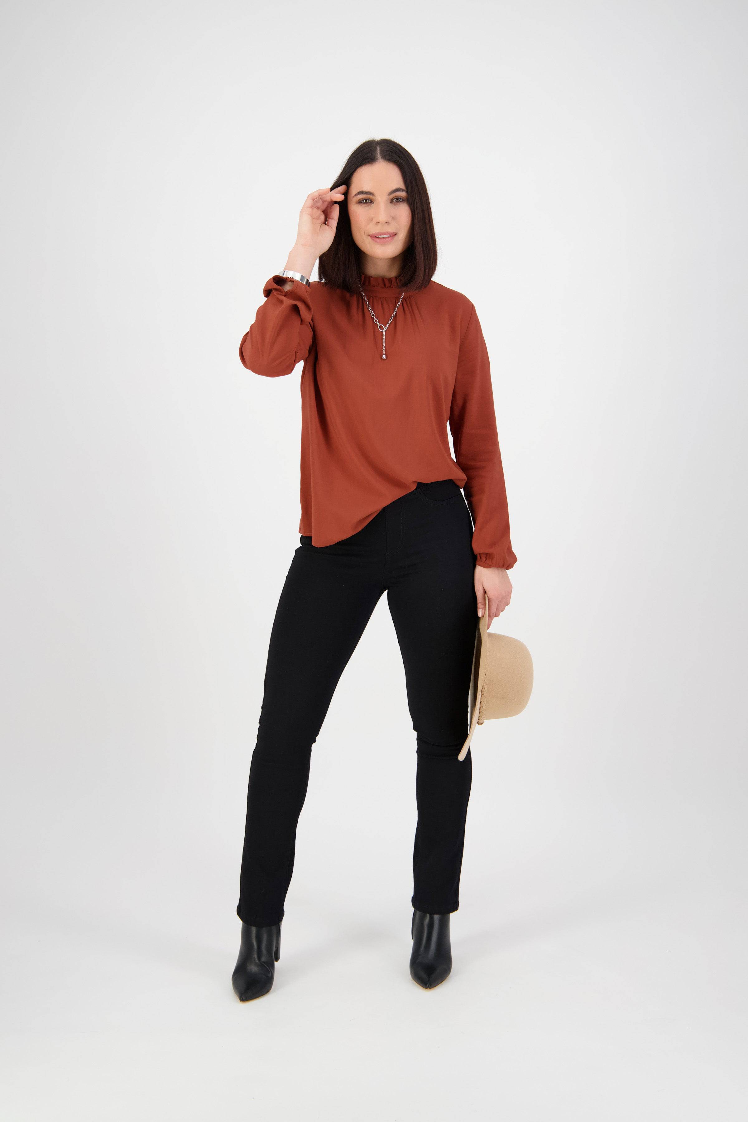 A woman stands against a plain white background, wearing a rust blouse, Vassalli Bay Knit Pull On - Black pants, black ankle boots, and holding a beige hat in her left hand.