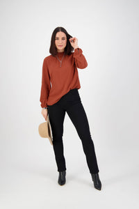A woman with shoulder-length dark hair in a rust blouse, Vassalli Bay Knit Pull On - Black pants, and black boots stands against a white background, holding a beige hat in one hand and touching her hair with the other.