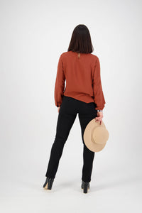 A woman faces away from the camera, wearing a rust long-sleeve blouse, Vassalli Bay Knit Pull On pants in black, black heeled shoes, and holding a beige hat in her right hand against a plain white background.