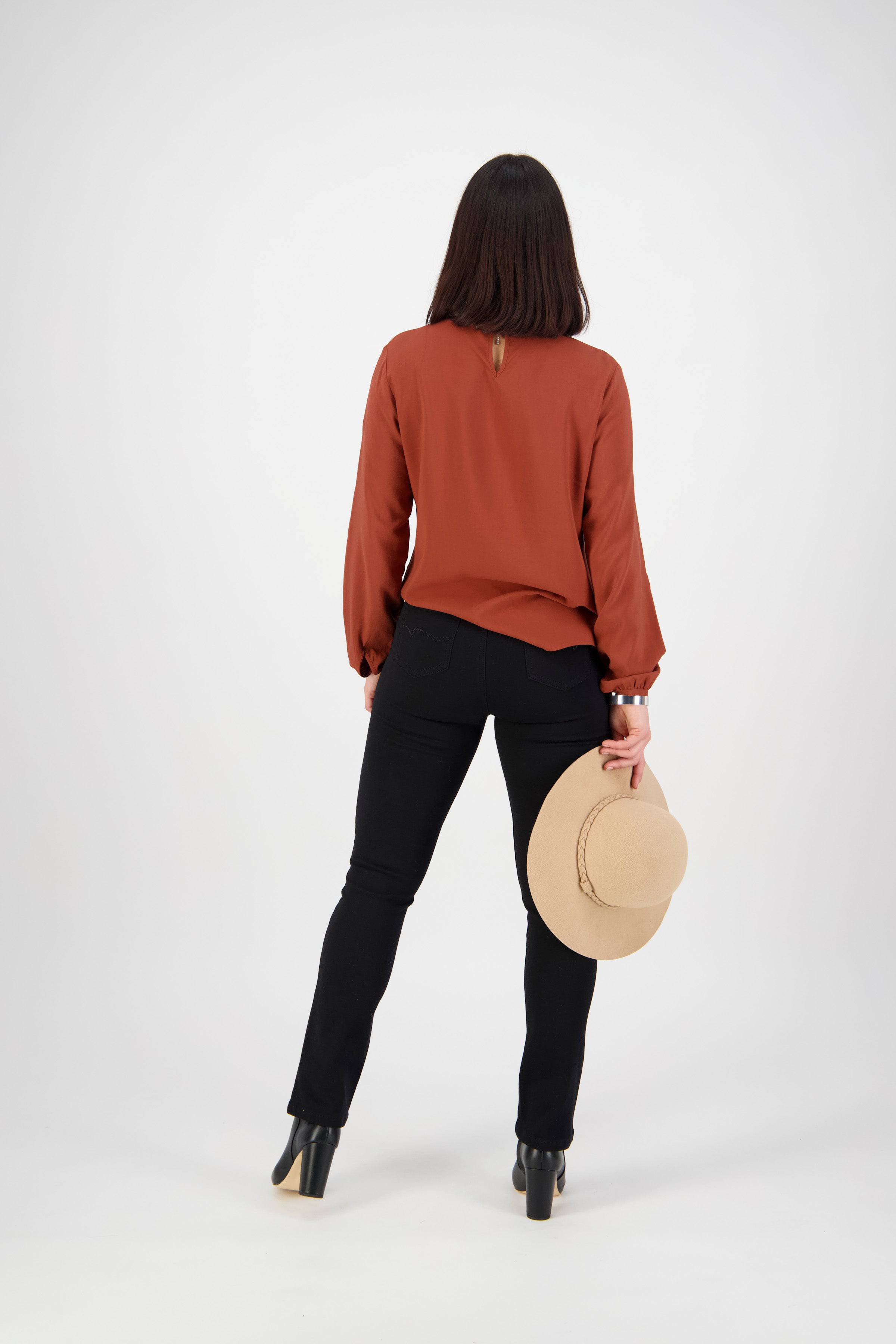 A woman faces away from the camera, wearing a rust long-sleeve blouse, Vassalli Bay Knit Pull On pants in black, black heeled shoes, and holding a beige hat in her right hand against a plain white background.
