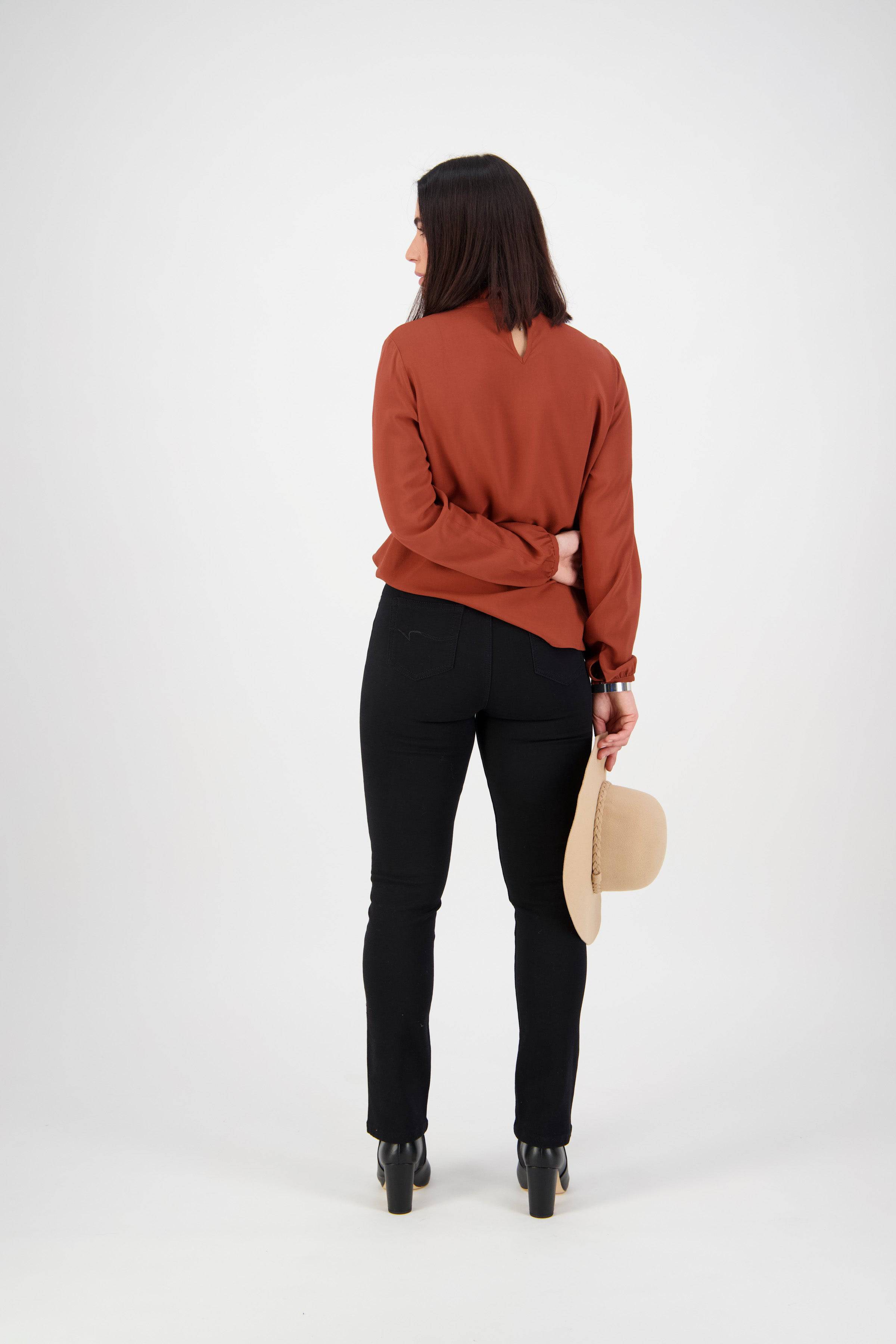 A woman with shoulder-length dark hair, wearing a rust blouse and Vassalli Bay Knit Pull On - Black pants, stands facing away from the camera and holds a beige wide-brimmed hat in her right hand.
