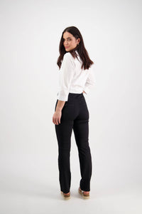 A woman with long dark hair wears the Vassalli Bay Pull On - Black slim leg pant, crafted from stretch fabric, and a white blouse. She stands against a plain white background, turned away but looking back with a smile.