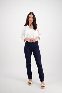A woman stands against a plain white background, wearing Vassalli's Bay Pull On - Navy slim leg pants with a white button-up shirt and platform sandals. She smiles warmly, her long dark hair loose and her hands lightly clasped.