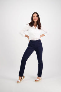 A woman with long dark hair stands confidently with hands on hips, wearing the Vassalli Bay Pull On - Navy slim leg pant in comfortable stretch fabric, paired with a white shirt and wedge sandals, against a plain white background.