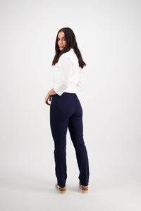 A woman with long dark hair, wearing a white shirt and Vassalli Bay Pull On pants in navy, stands turned slightly away and looks back over her shoulder against a plain white background.