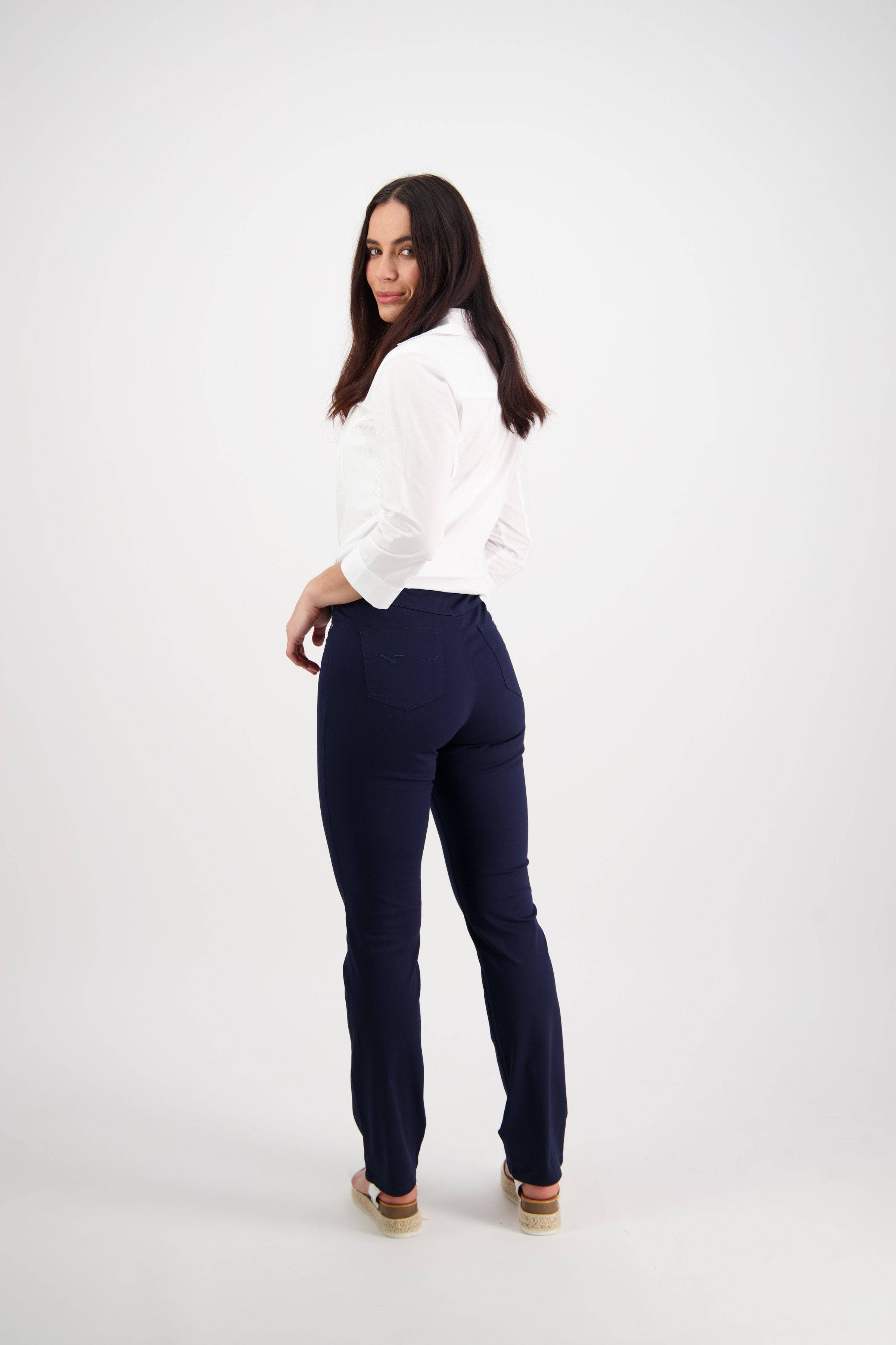A woman with long dark hair, wearing a white shirt and Vassalli Bay Pull On pants in navy, stands turned slightly away and looks back over her shoulder against a plain white background.