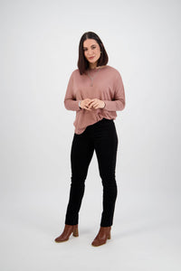 A woman with straight, shoulder-length brown hair stands against a white background, wearing a mauve long-sleeve top and Vassalli Bay Cord Pull On pants in black. She is smiling softly with her hands clasped in front of her.