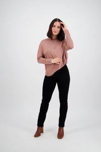 A woman with straight dark hair stands against a white background, wearing a mauve long-sleeve top, Vassalli Bay Cord Pull On pants in black, brown ankle boots, and a necklace. She is touching her hair and looking at the camera.