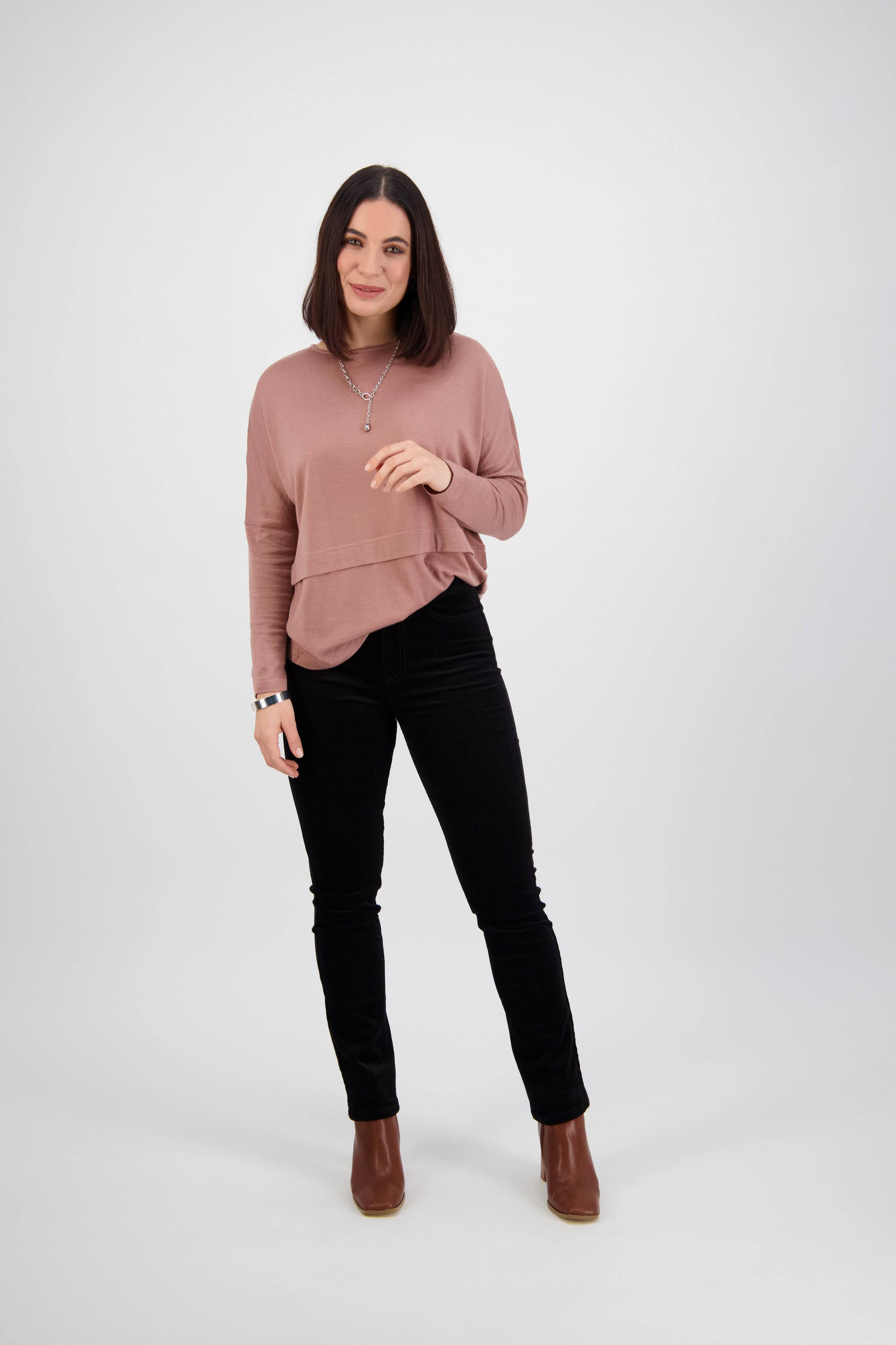 A woman with shoulder-length dark hair stands against a white background, wearing a mauve long-sleeve top, Vassalli Bay Cord Pull On pants in black, brown ankle boots, and a necklace, smiling with one hand near her face.