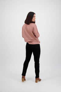 A woman with shoulder-length dark hair turns to smile at the camera, wearing a pink sweater, Vassalli Bay Cord Pull On pants in black, and brown heeled shoes against a plain white background.