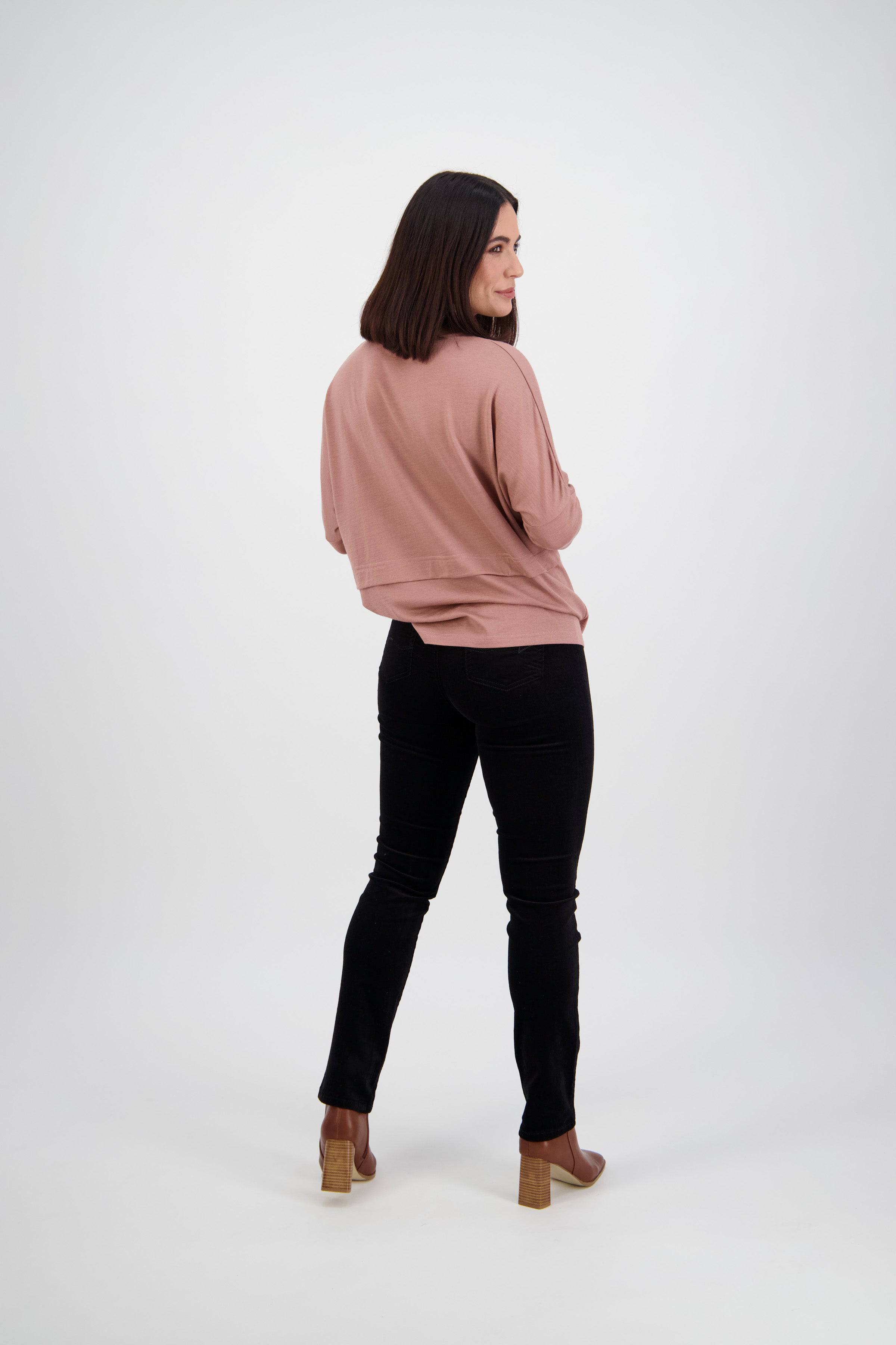A woman with shoulder-length dark hair turns to smile at the camera, wearing a pink sweater, Vassalli Bay Cord Pull On pants in black, and brown heeled shoes against a plain white background.