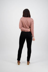 A person with shoulder-length brown hair, facing away, wears a pink long-sleeve top, Vassalli Bay Cord Pull On pants in black, and brown heeled shoes. One arm is bent behind their back against a plain white background.