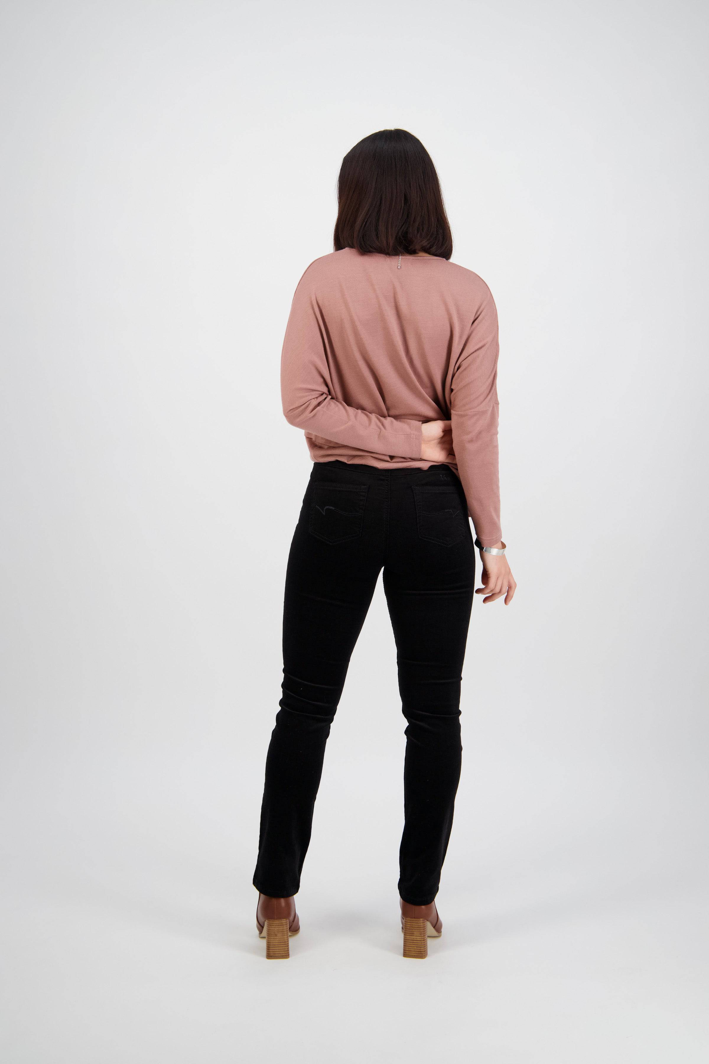 A person with shoulder-length brown hair, facing away, wears a pink long-sleeve top, Vassalli Bay Cord Pull On pants in black, and brown heeled shoes. One arm is bent behind their back against a plain white background.